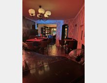 Eine gemütliche Bar mit gedimmter Beleuchtung, Tapeten mit floralem Muster und Holzböden. Im Raum befinden sich hohe Tische mit Barhockern, ein Kronleuchter mit runden Lampenschirmen und eine Theke im Hintergrund mit beleuchteten Regalen voller Getränke.