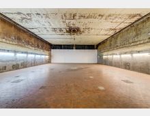 Ehemaliger industrieller Raum mit hohen Betonwänden und abgenutzter Decke, beleuchtet durch eine Reihe von linearen Lichtquellen an den Seiten. Der Boden besteht aus rechteckigen, rötlichen Fliesen, und die Umgebung wirkt roh und unverkleidet.