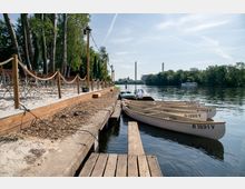 Ein Uferbereich mit befestigtem Holzsteg, an dem weiße Ruderboote vertäut sind. Links befinden sich ein sandiger Gehweg, Holzgeländer und aufgestellte Tische unter Bäumen; im Hintergrund führt ein Fluss mit bewaldeten Ufern zu einer Skyline mit Industrieschornsteinen.
