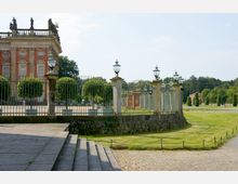 Teilansicht eines historischen Gebäudes mit roter Fassade und weißen Fenstern, flankiert von Steinpfeilern mit dekorativen Laternen und einem Eisengitterzaun. Im Vordergrund sind gepflasterte Wege und Treppenstufen, im Hintergrund ein parkähnliches Gelände mit Bäumen und grasbewachsenen Flächen.