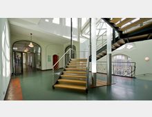 Modernes Treppenhaus in einem Gebäude mit geradliniger Holztreppe und Metallgeländer, umgeben von großen Fenstern, die Tageslicht hereinlassen. Im Hintergrund sind ein Gang mit Glastüren und geschwungene architektonische Details sichtbar.