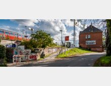 Bahnhofsumgebung mit einer Unterführung, deren Wände mit Graffiti bedeckt sind, einem roten Backsteingebäude auf der rechten Seite und einem Zuggleis im oberen Bereich, auf dem ein roter Zug sichtbar ist. Ein Schild mit der Aufschrift „Zossen“ markiert den Standort, und die Szene wird von Bäumen und einer asphaltierten Straße eingerahmt.