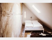 Minimalistisch eingerichtetes Schlafzimmer in einem Dachgeschoss mit schräger, weißer Decke. Das Zimmer zeigt ein weißes Doppelbett, Holzfußboden, eine schmale Wandöffnung als Fenster und eine braune Fellteppich-Dekoration.