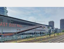 Außenansicht der "East Side Mall", einem modernen Einkaufszentrum mit horizontalen Lamellenfassaden und orangefarbener Beschriftung. Im Vordergrund befinden sich Eisenbahngleise, Signalpfosten und eine mit Graffiti besprühte Betonwand; im Hintergrund sind zwei Hochhäuser und ein Kran zu sehen.