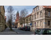 Straßenszene in einer historischen Stadt mit gepflastertem Straßenbelag, flankiert von alten Gebäuden mit teilweise verwitterter Fassade. Im Hintergrund ragt ein roter Kirchturm mit grünem Spitzdach empor, während geparkte Autos entlang der Straße stehen.
