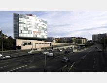 Modernes Bürogebäude mit auffälliger Glasfassade, das auf einem sandfarbenen Unterbau ruht, neben einer mehrspurigen Straße mit fließendem Verkehr; im Hintergrund sind Wohnblocks und Bäume zu sehen.