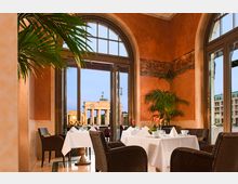 Innenansicht eines eleganten Restaurants mit großen Rundbogenfenstern, die einen Blick auf das Brandenburger Tor bieten. Der Raum ist mit orangen Wänden, Pflanzen und gedeckten Tischen mit weißen Tischdecken gestaltet.