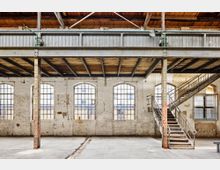 Industriehalle mit hohen, weiß gestrichenen Ziegelwänden und großen, bogenförmigen Fenstern. Im Vordergrund sind Metallstützen, eine Betondecke und eine Treppe mit Metallgeländer zu sehen, während Holzbalken die Decke bilden.
