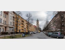 Straßenszene in einer Stadt mit mehrstöckigen Wohngebäuden auf beiden Seiten, teilweise in gelber und beigefarbener Fassade. Im Hintergrund steht ein Kirchturm mit grünlicher Kuppel, während Autos entlang der Straße und geparkte Fahrräder am Gehweg zu sehen sind.