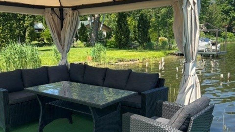 Überdachte Lounge-Ecke auf einer grünen Plattform am Wasser mit Rattan-Sofaecke, Sesseln und Glastisch unter einem Pavillon mit Vorhängen. Im Hintergrund sind ein ruhiger Teich oder Kanal, Uferbewuchs und Bäume zu sehen.