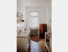 Helle, schmale Küche mit Holzboden, einer weißen Arbeitsplatte mit Waschbecken auf der linken Seite, einem roten Vintage-Kühlschrank und einem Herd rechts. Ein großes Fenster mit darunterliegendem Heizkörper erhellt den Raum, vor dem ein roter Teppich liegt.