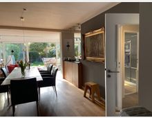 Wohnzimmer mit offenem Essbereich und großen bodentiefen Fenstern, die auf einen Garten mit Terrasse blicken. Der Raum hat einen Holzboden, einen Esstisch mit schwarzen Stühlen und eine schlichte Einrichtung mit dekorativen Elementen wie einem Gemälde an der Wand.