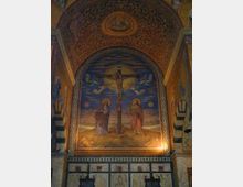 Innenraum einer Kirche mit einem Fresko, das die Kreuzigung Christi darstellt. Das Kunstwerk ist von reich verzierten, goldenen und floralen Mustern umrahmt, die sich über die Wölbung der Apsis erstrecken.