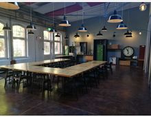 Ein industriell gestalteter Raum mit großen Fenstern, mehreren rechteckigen Holztischen, schwarzen Stühlen und zahlreichen hängenden Lampen. Im Hintergrund befinden sich Regale, ein großer Wandwecker und Dekorationselemente, die den Raum auflockern.