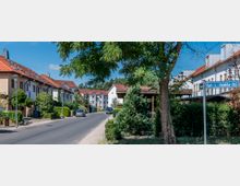 Wohnstraße mit Reihenhäusern in einer Wohnsiedlung, gekennzeichnet durch weiße Fassaden und rote Ziegeldächer. Im Vordergrund befindet sich ein Straßenschild („Carl-v.-Ossietzky-Str.“), umgeben von Bäumen, Hecken und parkenden Autos.