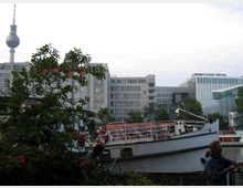 Städtische Szene mit einem weißen Boot am Wasser im Vordergrund, umgeben von Pflanzen. Im Hintergrund sind moderne Bürogebäude und der Berliner Fernsehturm sichtbar.