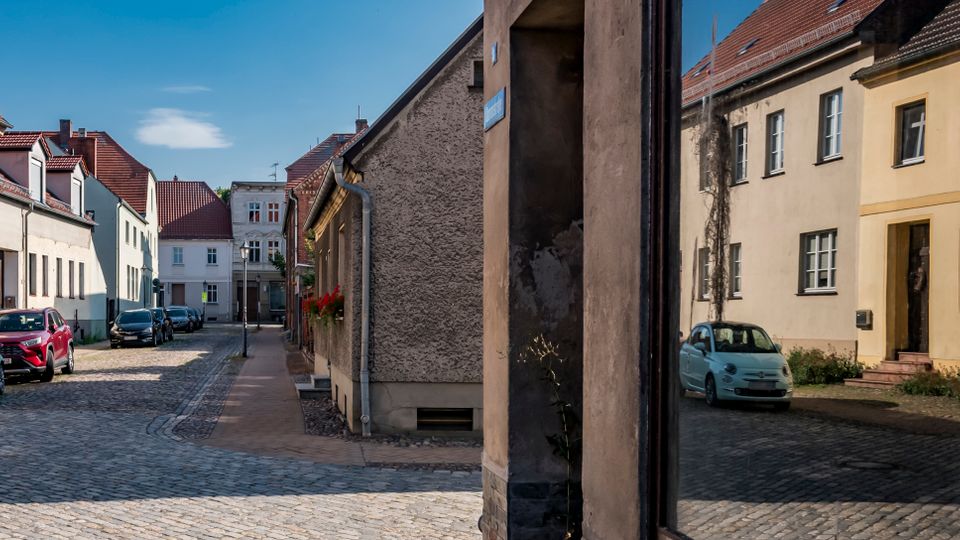 Blick auf eine schmale, gepflasterte Altstadtstraße mit niedrigen Wohnhäusern in hellen Fassaden und roten Ziegeldächern. Am rechten Bildrand steht eine spiegelnde Glasfläche, die die gegenüberliegenden Gebäude und geparkte Autos reflektiert.