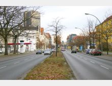 Breite, mehrspurige Stadtstraße mit Autos in beiden Fahrtrichtungen und einer begrünten Mittelinsel. Am Rand der Straße stehen unbelaubte Bäume, Wohn- und Geschäftsgebäude sowie eine Tankstelle; im Hintergrund ist ein modernes Hochhaus sichtbar.