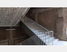 Betontreppe in einem Industriegebäude mit unverputzten Ziegelwänden und sichtbarer Deckenkonstruktion aus Beton. Das Geländer der Treppe ist aus Metall gefertigt, und die Umgebung vermittelt einen rohen, unfertigen Charakter.