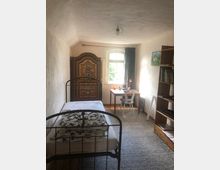 Schmaler Raum mit schlichtem Interieur: ein schmiedeeisernes Einzelbett, ein verziertes, bunt bemaltes Holzschrankelement, ein Schreibtisch mit Stuhl vor einem Fenster mit halb geöffnetem Vorhang sowie ein Holzregal an der rechten Wand.