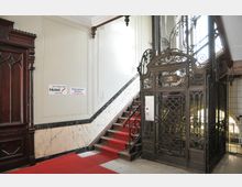 Treppenhaus mit einem historischen Aufzug aus schwarz verziertem Metall neben einer Treppe mit rotem Teppich. Die Wände sind teils mit Marmor verkleidet, und Hinweisschilder informieren über ein Hotel und eine Zahnarztpraxis in den oberen Etagen.