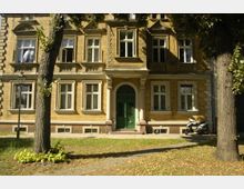 Mehrstöckiges Wohngebäude aus gelbem Klinkerstein mit symmetrisch angeordneten weißen Fenstern und einem grünen Eingangsportal. Im Vordergrund stehen zwei Bäume, ein Weg führt zum Eingang, und ein Roller ist an der rechten Seite geparkt.