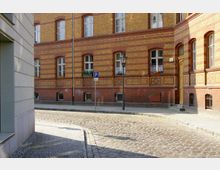 Straßenszene mit einem historischen Gebäude aus gelb-roten Ziegeln, Fenstern mit weißen Rahmen und Blumentöpfen auf den Fensterbänken; gepflasterte Straße und Gehweg aus Kopfsteinpflaster, ein Laternenmast und ein Parkverbotsschild sind sichtbar.