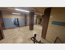 Ein Innenraum mit gefliesten Wänden, bei denen blaue und beige Fliesen kombiniert sind, sowie Backsteinpfeilern. Der Boden besteht aus beigefarbenen Fliesen, eine Treppe mit Metallgeländer führt in den Raum, und an der Wand ist ein Schild mit der Aufschrift „Keine Schwimmhalle“ angebracht.