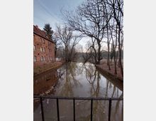 Backsteinbau neben einem ruhigen Fluss in einer von kahlen Bäumen gesäumten Landschaft; ein dunkles Geländer im Vordergrund und ein wolkenloser Himmel im Hintergrund.