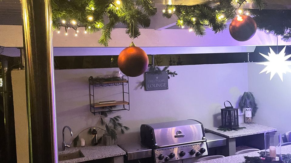 Außenküche mit überdachtem Bereich, geschmückt mit Tannengirlanden, Lichterketten und Kugeln. Ausstattung umfasst eine Stein-Arbeitsplatte, Edelstahlgrill, Spüle und dekorative Elemente wie Laternen und Pflanzen.