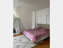 Schlafzimmer mit minimalistischem Design, ausgestattet mit einem Doppelbett mit rosa Bettwäsche, einem hellen Teppich auf Holzfußboden und einem großen Spiegel an der Wand. Eine runde, weiße Papierlampe hängt von der Decke, die Wände sind schlicht weiß gestrichen.