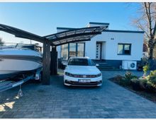 Moderne, einstöckige weiße Wohnhausfassade mit einem flachen Dach und großen Fenstern, davor ein gepflasterter Hof mit einem überdachten Stellplatz, auf dem ein Auto und ein Boot auf einem Anhänger parken. Im Vordergrund sind dekorative Pflanzen und eine Wärmepumpe an der Seite des Hauses sichtbar.