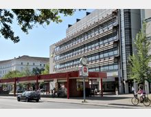 Moderne Straßenansicht mit einem mehrstöckigen Bürogebäude mit Glas- und Metallfassade im Hintergrund. Im Vordergrund befindet sich eine rote Überdachung mit einer Uhr und Werbeschildern sowie Fußgänger und Fahrradfahrer auf dem Gehweg.