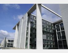 Modernes Gebäude mit klaren Linien aus Beton und Glasfassaden, das durch große Fensterflächen und offene rechteckige Strukturen geprägt ist; im Hintergrund blauer Himmel mit vereinzelten Wolken.