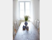Ein helles Esszimmer mit einem rustikalen Holztisch, umgeben von weißen Holzstühlen. Auf dem Tisch stehen eine blau-weiße Vase mit Blumen sowie kleine, dekorative Keramikgefäße. Im Hintergrund befindet sich ein großes Fenster mit weißen, lichtdurchlässigen Vorhängen.