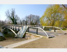 Moderne Fußgängerbrücke mit geschwungenen Geländern aus Metall und Holzbelag, die über einen kleinen Fluss führt. Im Hintergrund stehen blattlose Bäume und eine große Trauerweide, umgeben von einem parkähnlichen Gelände.