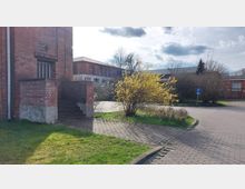 Außenansicht eines Ziegelgebäudes mit einer Außentreppe, umgeben von einem gepflasterten Weg, einem gelb blühenden Strauch und weiteren niedrigen Büschen. Im Hintergrund sind ein weiteres Ziegelgebäude, ein Parkplatz und vereinzelt Bäume sowie ein Schild mit einem "P"-Symbol erkennbar.