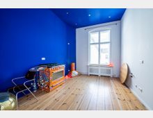 Blauer Raum mit Holzdielenboden und einer großen Fensterfront auf der rechten Seite. Links stehen ein dekorativer, bunt bemalter Herd, ein moderner Stuhl, eine Discokugel und bunte Lichter; an der Wand hängt leuchtende Dekoration.