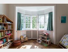 Ein Kinderzimmer mit einer großen, halbkreisförmigen Fensterfront, die von grünen Vorhängen eingerahmt ist. Links steht ein Holzregal mit Büchern und Spielzeug, rechts ein Puppenhaus auf dem Boden.