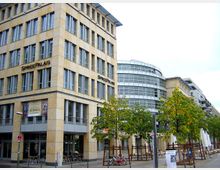 Modernes Bürogebäude namens "Spreepalais" mit beigefarbener Fassade und großen Fenstern, daneben ein rundliches, verglastes Gebäude. Im Vordergrund sind Bäume, Fahrradständer und Sitzbereiche mit Tischen und Sonnenschirmen zu sehen.