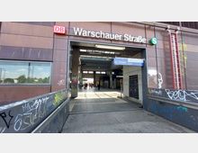 Eingangsbereich des S- und Regionalbahnhofs Warschauer Straße in Berlin, umgeben von einer modernen Fassade mit Graffiti. Das Gebäude besitzt offene Türen, durch die das Innere mit Glasdächern erkennbar ist, sowie ein Schild mit Wegweisern zu den Zügen.