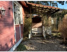 Eine Holzwand eines rosa gestrichenen Gebäudes mit einem vergitterten Fenster auf der linken Seite. Rechts befindet sich ein kleiner überdachter Eingang mit einer Pergola, umgeben von karger Vegetation und Kiesboden.