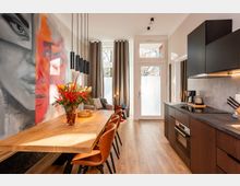 Moderner, schmaler Küchen- und Essbereich mit langem Holztisch, braunen Stühlen und schwarzen Pendelleuchten sowie einer Küchenzeile mit dunklen Schränken und grauer Arbeitsplatte. An der linken Wand befindet sich ein großformatiges Wandgemälde, am Ende des Raums führen hohe Fenster und eine Glastür mit Vorhängen nach außen.