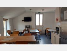 Ein Gemeinschaftsraum mit mehreren Tischen und Holzstühlen, einer Küchenzeile, einem Kühlschrank, einer Mikrowelle und einem an der Wand montierten Fernseher; die Wände sind weiß, mit einem Fenster und einer Glastür, die Tageslicht hereinlassen.