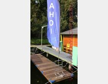 Ein kleiner Steg aus Holz mit blauen Kantenschonern, befestigt an einem Metallsteg vor einer hölzernen, orangefarbenen Hütte im Grünen. Links im Bild steht eine blaue Fahne mit der Aufschrift „AHOI“.