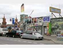 Straßenszene in Berlin mit einem Abschnitt der East Side Gallery, einer bemalten Betonmauer mit kunstvollen Graffitis, darunter ein Soldat und historische Szenen. Links ist die Oberbaumbrücke mit ihren markanten Türmen zu sehen. Im Vordergrund stehen geparkte Autos und ein Souvenirgeschäft mit einer deutschen Flagge auf dem Dach.