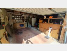 Eine geöffnete Garage mit Werkstattcharakter, ausgestattet mit Regalen, Werkzeugen, Materialien und einer Holzwerkbank. Der Boden ist teilweise unaufgeräumt, und im Hintergrund sind Lagergegenstände wie Leitern und Kartons sichtbar.