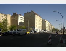 Stadtszene mit mehrstöckigen Wohngebäuden im klassischen Stil, geparkten und fahrenden Autos entlang einer breiten Straße. Im Vordergrund stehen Baustellenabsperrungen, und hohe Straßenlaternen säumen den Straßenrand unter einem klaren Himmel.