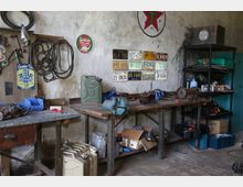 Eine Werkstatt mit alten Werkbänken, an denen Werkzeuge, Kanister und Autoteile liegen. An der Wand hängen Nummernschilder, Schilder und ein Texaco-Logo, daneben befindet sich ein Regal mit Werkzeugen und Zubehör.