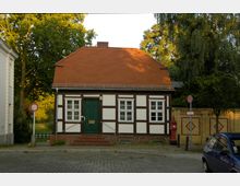 Fachwerkhaus mit weißer Fassade und dunklen Holzbalken, rotem Satteldach und grüner Holztür, umgeben von Bäumen und einer Ziegelmauer im Hintergrund. Im Vordergrund führt eine gepflasterte Straße vorbei.